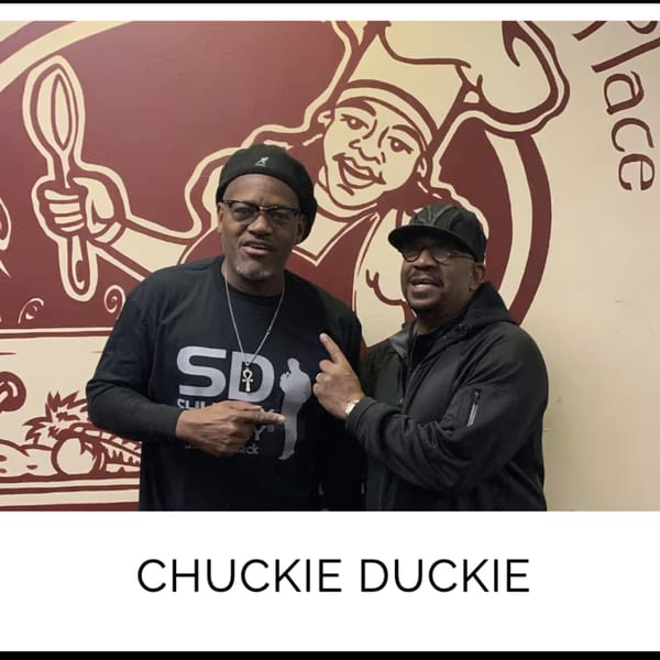 Two men, Chuckie Duckie, stand in front of a Kountry Kitchen chef mural, pointing at each other.