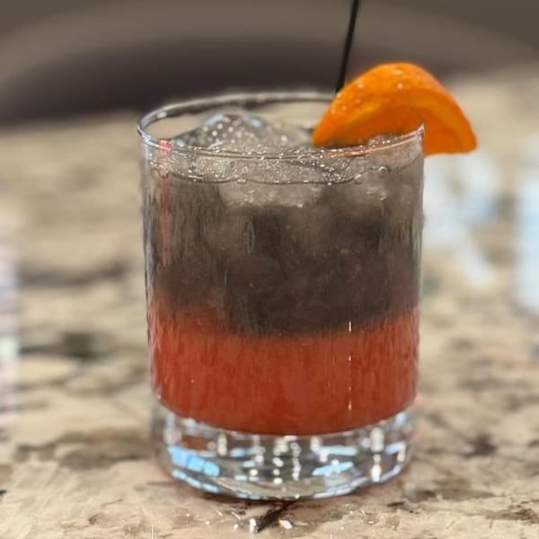 A layered cocktail with red and dark gray liquid, ice, and an orange slice garnish.
