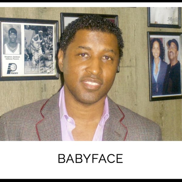 Babyface, a Black man in a plaid jacket and lavender shirt, smiles in front of framed photos.