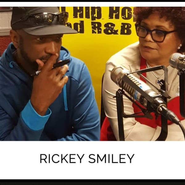 Two people, one in a blue hoodie and cap, the other in glasses, speaking into microphones.