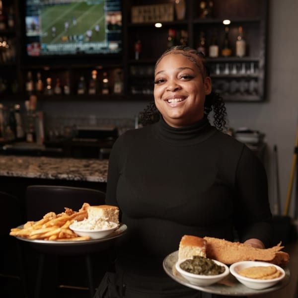 Smiling Kountry Kitchen server holding two trays of soul food: fried fish, fries, coleslaw, cornbread, and greens.